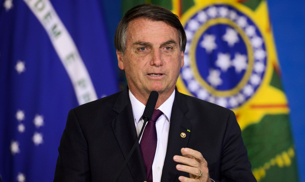 Jair Bolsonaro falando e gesticulando, com bandeira do Brasil ao fundo