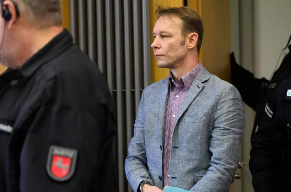 Christian Brueckner em pé em tribunal, com camisa roxa e paletó cinza, com expressão séria