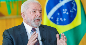 Lula falando e gesticulando com expressão séria e bandeira do Brasil ao fundo