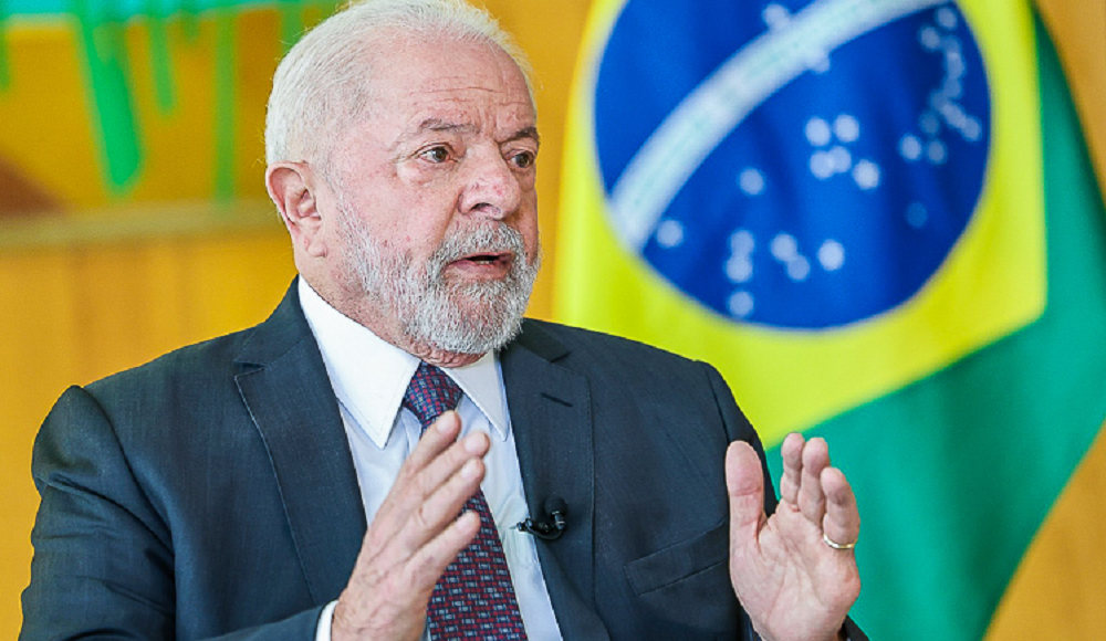 Lula falando e gesticulando com expressão séria e bandeira do Brasil ao fundo