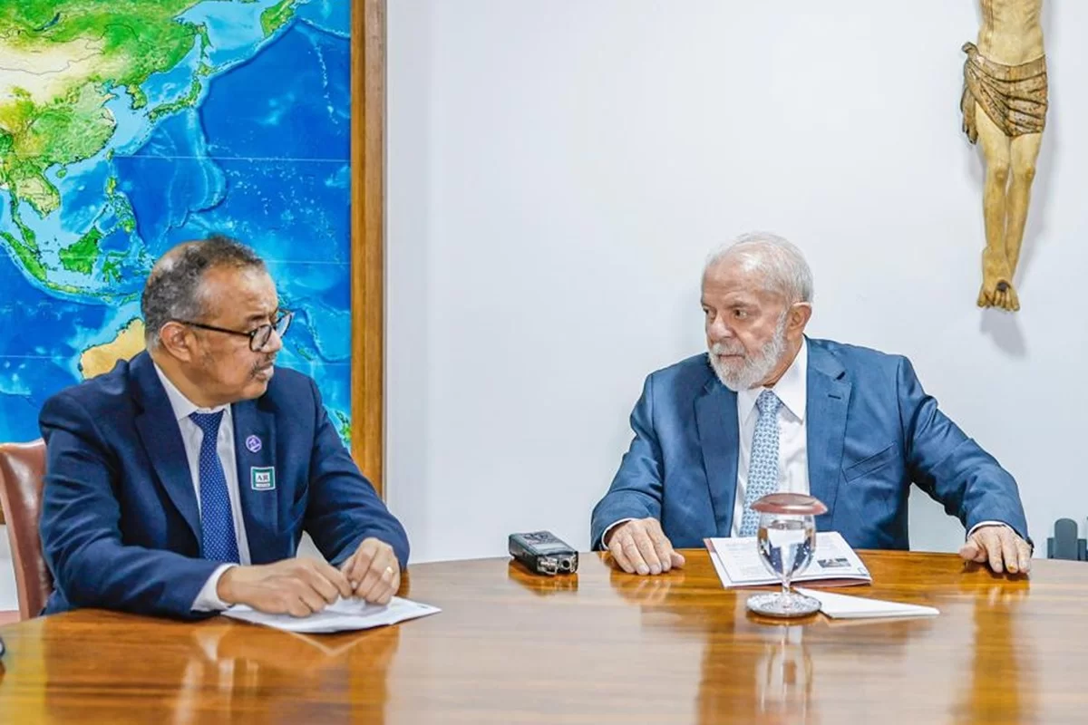 Tedros Adhanom e Lula sentados lado a lado, conversando com expressão séria