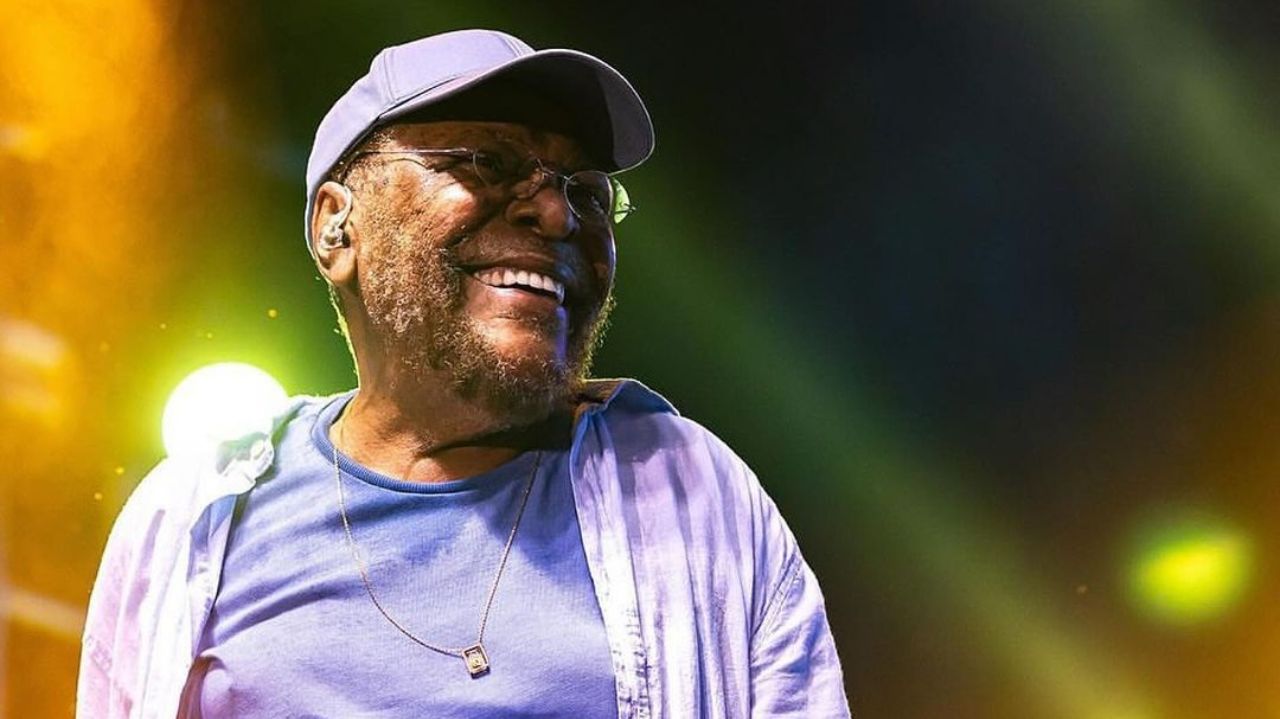 Martinho da Vila de roupa e boné azuis, com óculos, olhando para o lado e sorrindo