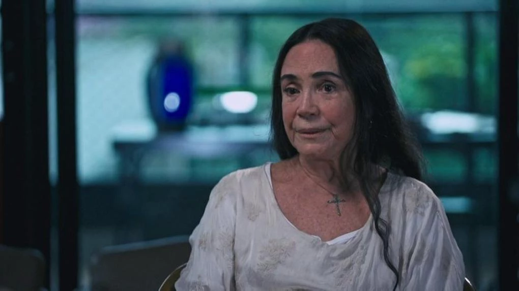 Regina Duarte de roupa clara e cabelo solto, falando com expressão séria