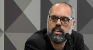 Allan dos Santos falando em microfone com expressão séria, de óculos