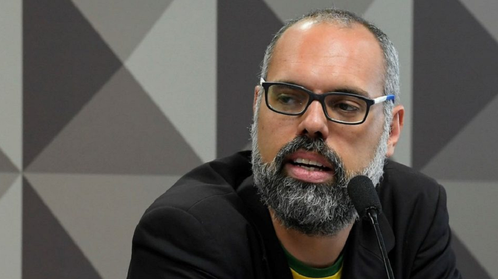 Allan dos Santos falando em microfone com expressão séria, de óculos