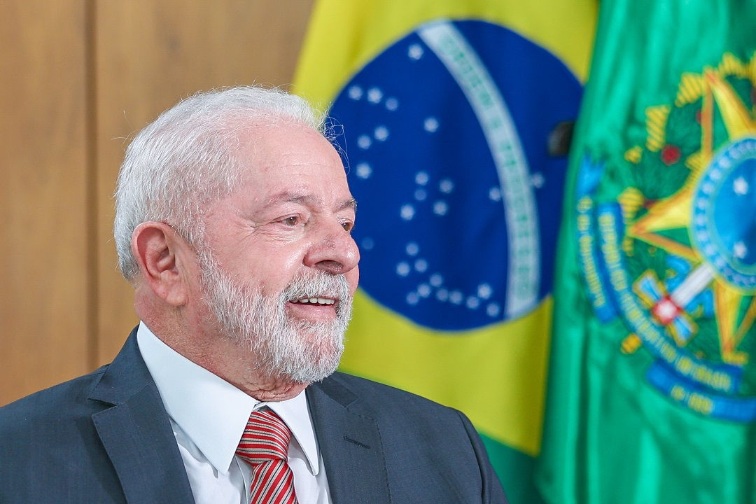 Lula de perfil, sorrindo, perto de bandeira brasileira