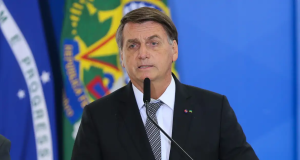 Jair Bolsonaro falando em microfone com expressão séria e bandeiras ao fundo