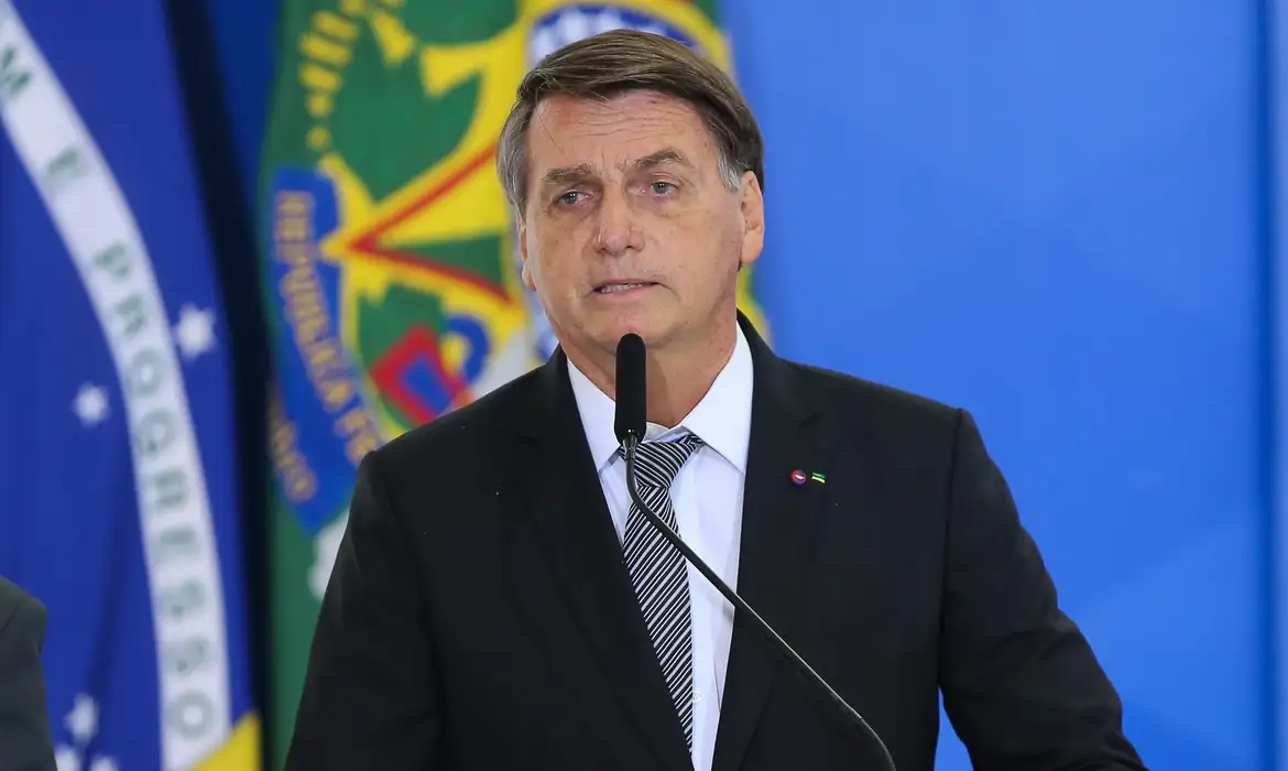 Jair Bolsonaro falando em microfone com expressão séria e bandeiras ao fundo