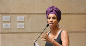 Marielle Franco de camiseta preta e turbante roxo, falando em microfone e gesticulando, com expressão séria