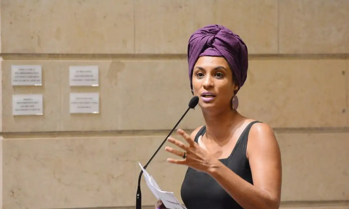 Marielle Franco de camiseta preta e turbante roxo, falando em microfone e gesticulando, com expressão séria