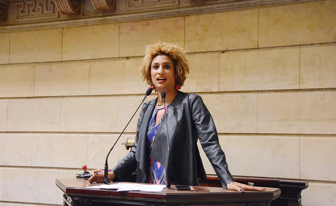 Marielle Franco em pé, falando em microfone com expressão séria
