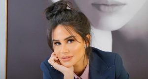 Natalia Martins posando de roupa social azul e rosa, com cabelo preso e rosto apoiado na mão