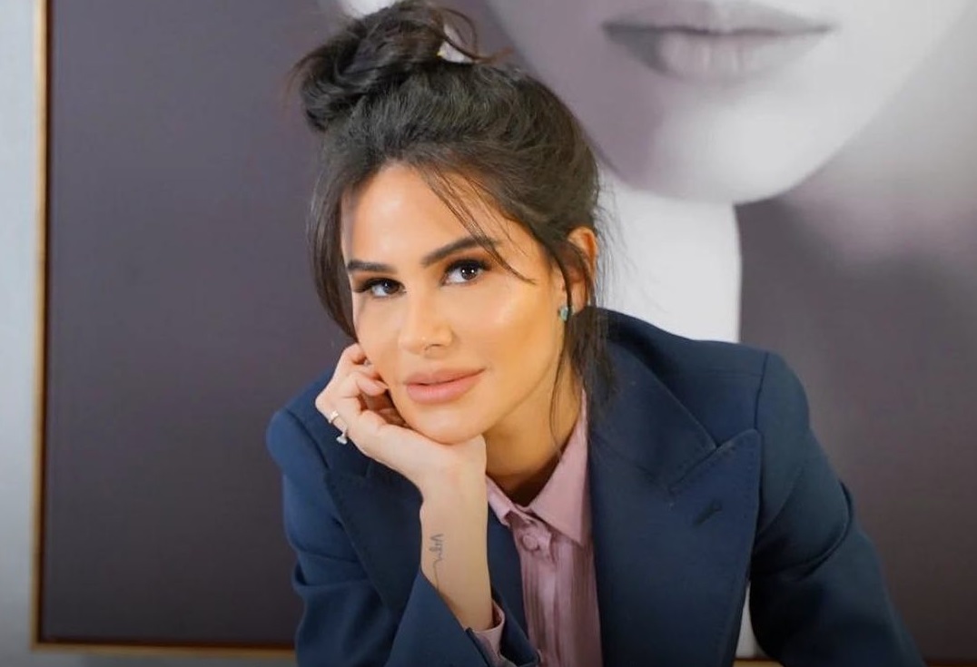 Natalia Martins posando de roupa social azul e rosa, com cabelo preso e rosto apoiado na mão