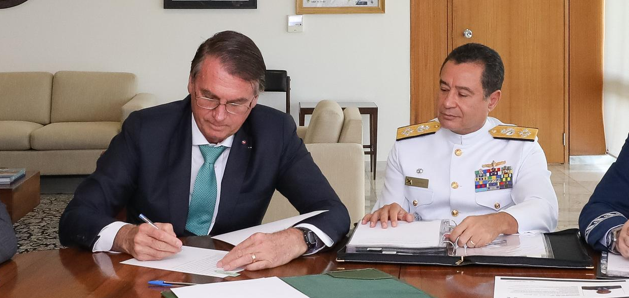 Jair Bolsonaro assinando documento e sendo observado por Almir Garnier Santos