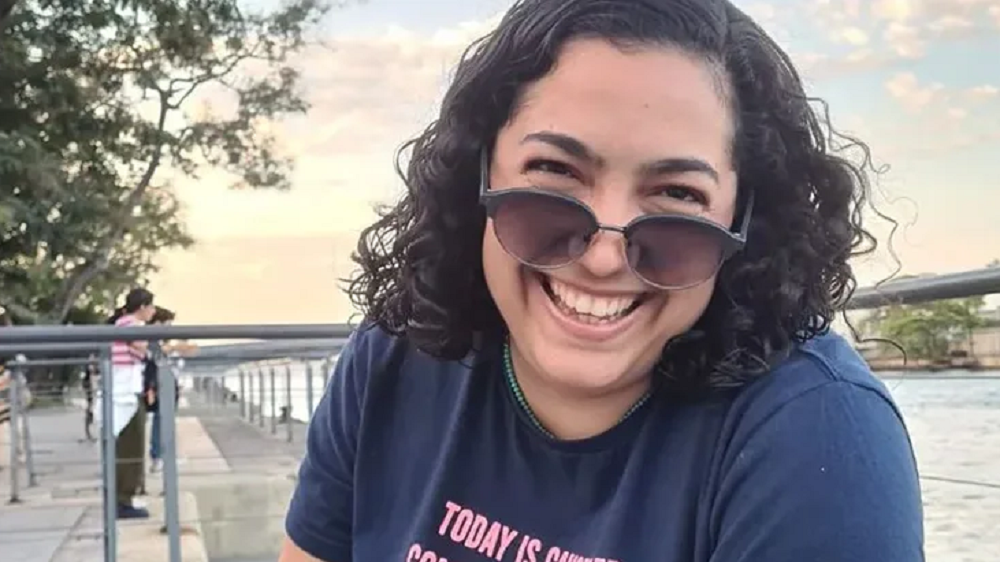 Camila Moura de camiseta azul e óculos escuros, sorrindo para a câmera