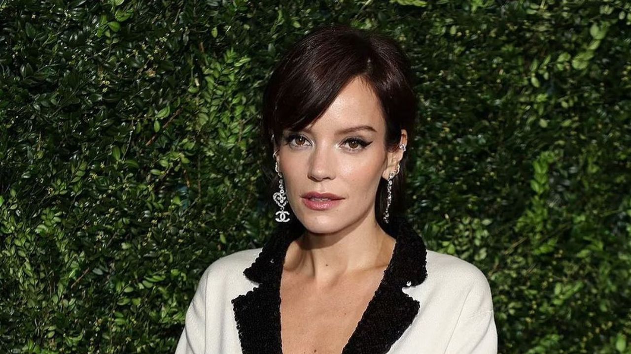 Lily Allen com roupa preta e branca, fazendo carão, perto de parede de plantas