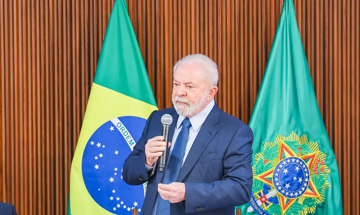 Lula falando em microfone com bandeiras ao fundo, com expressão séria
