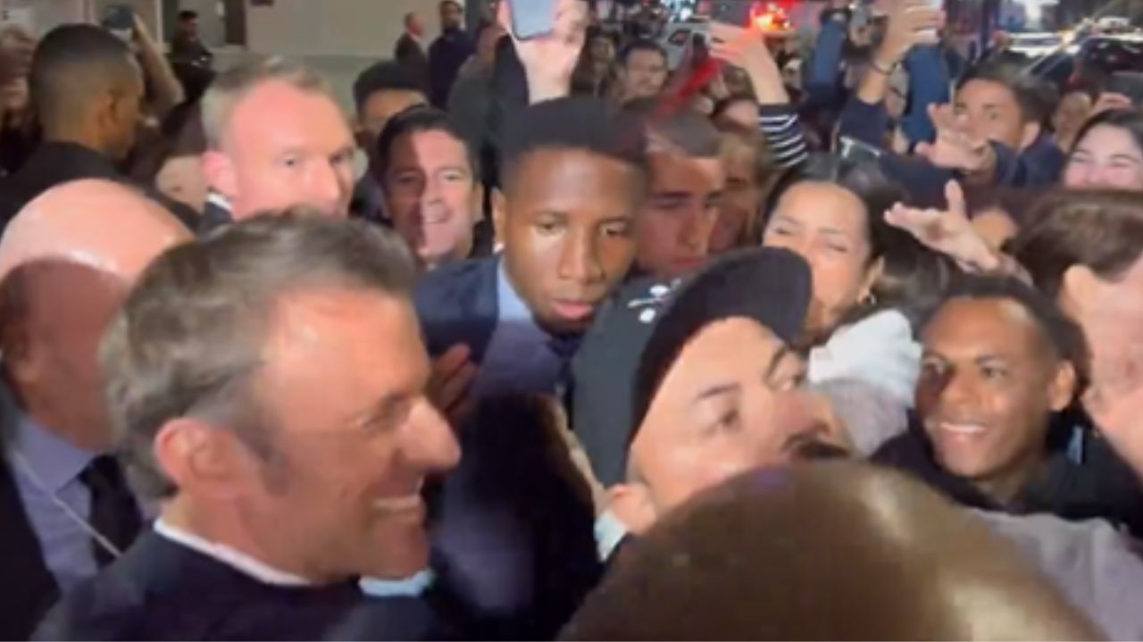 Emmanuel Macron cercado por pessoas, sorrindo, de perfil