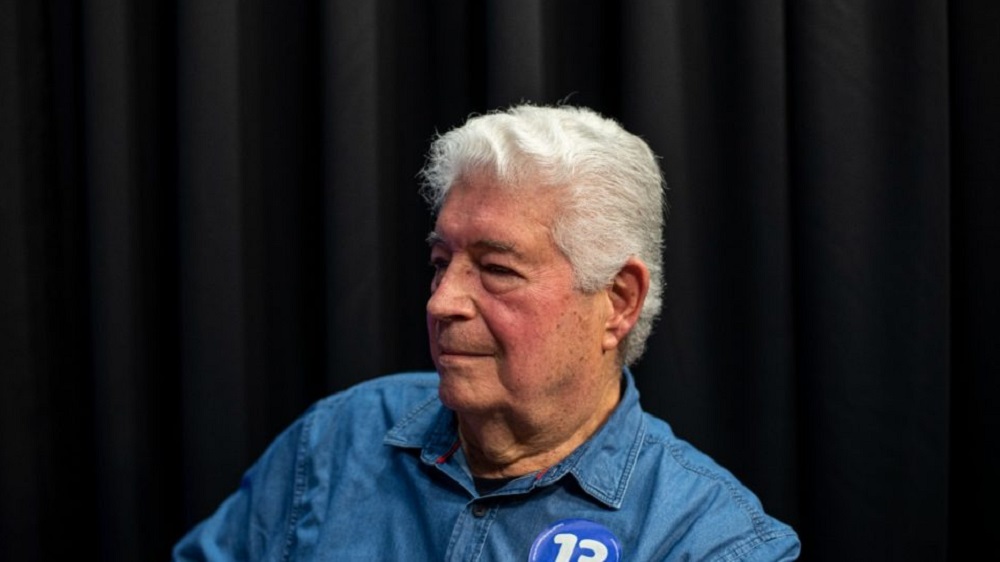 Roberto Requião sério, de perfil, com camisa jeans e adesivo escrito 13