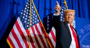 Donald Trump com expressão de satisfação, apontando pra cima perto de bandeira dos EUA