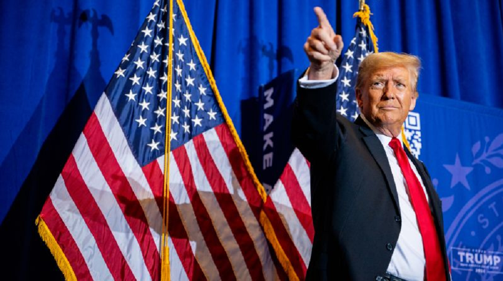 Donald Trump com expressão de satisfação, apontando pra cima perto de bandeira dos EUA