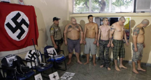 Grupo de homens sem camisa em pé, perto de agente e de símbolos nazistas