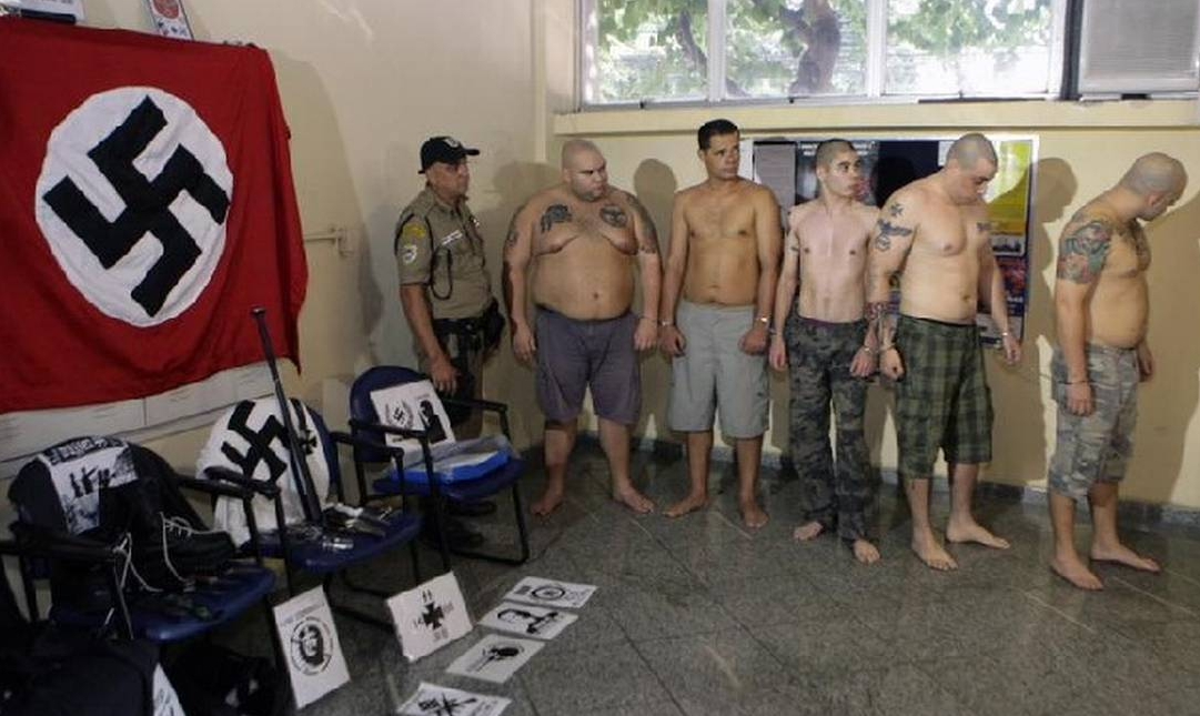 Grupo de homens sem camisa em pé, perto de agente e de símbolos nazistas