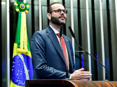 Jorge Seif de óculos, falando em microfone com bandeira do Brasil ao fundo