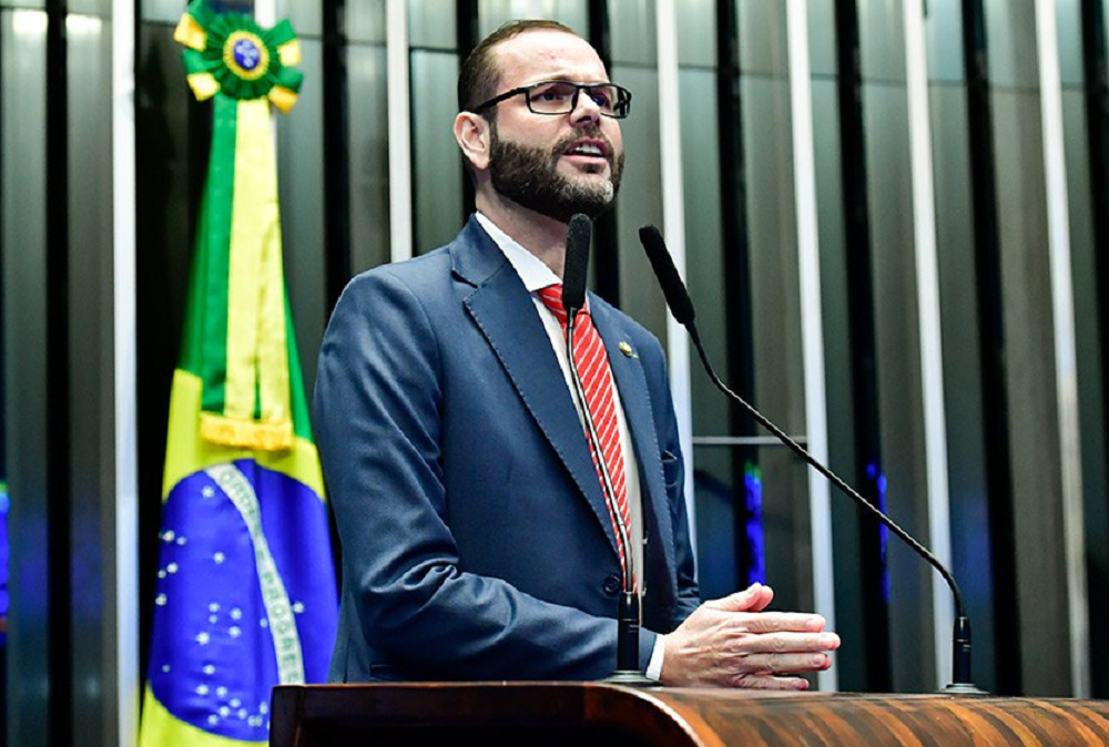 Jorge Seif de óculos, falando em microfone com bandeira do Brasil ao fundo