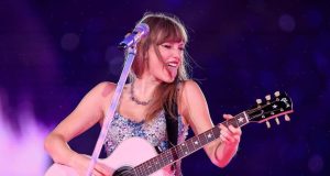 Taylor Swift no palco, tocando violão e fazendo careta com a língua de fora