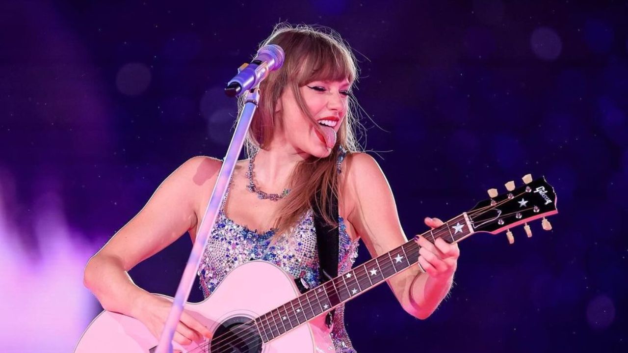 Taylor Swift no palco, tocando violão e fazendo careta com a língua de fora