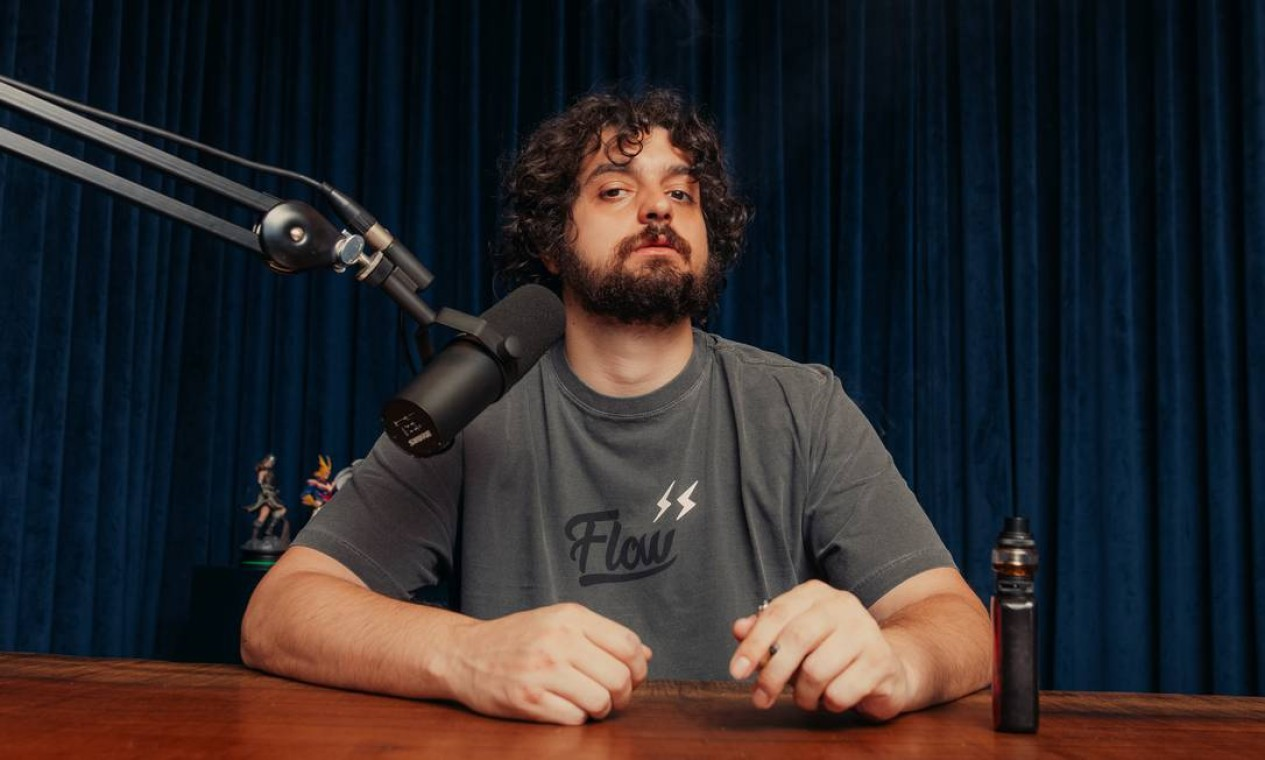 Monark posando para foto em estúdio, com expressão séria e camiseta cinza do Flow