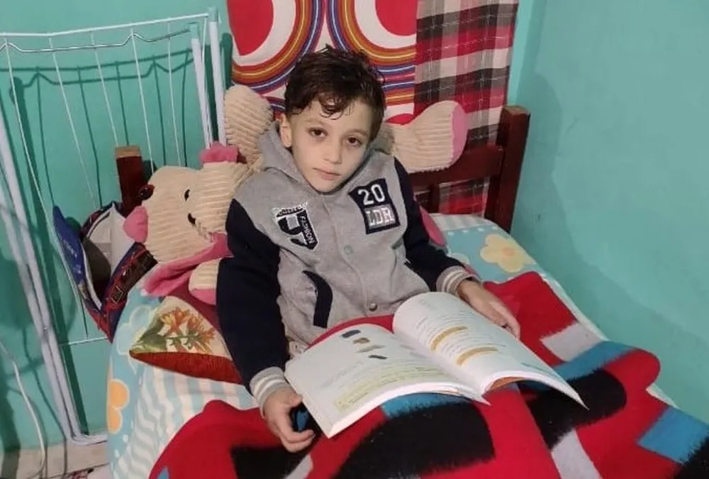 Miguel olhando para a câmera, sério, deitado em cama, segurando livro aberto