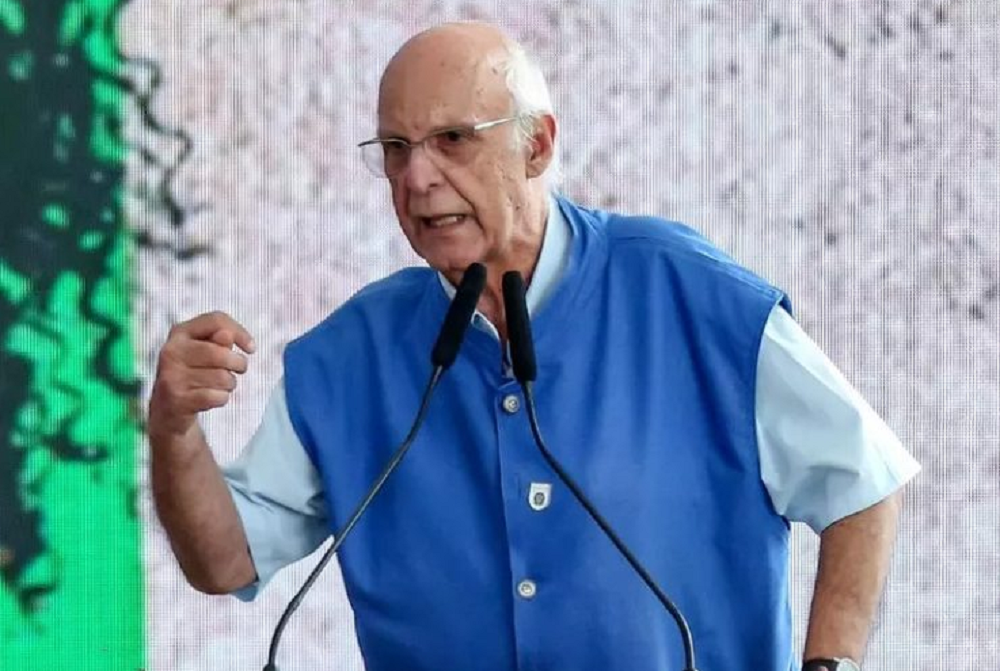 Padre Júlio Lancellotti de roupa branca e azul, falando em microfone e gesticulando com expressão indignada