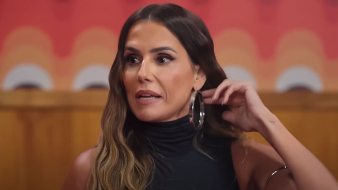 Deborah Secco de cabelo solto, camiseta preta e brincos de argola, falando e olhando para o lado