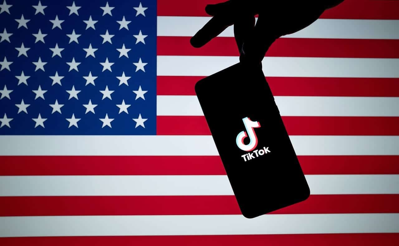 Mão segurando celular com logo do TikTok com bandeira dos EUA ao fundo