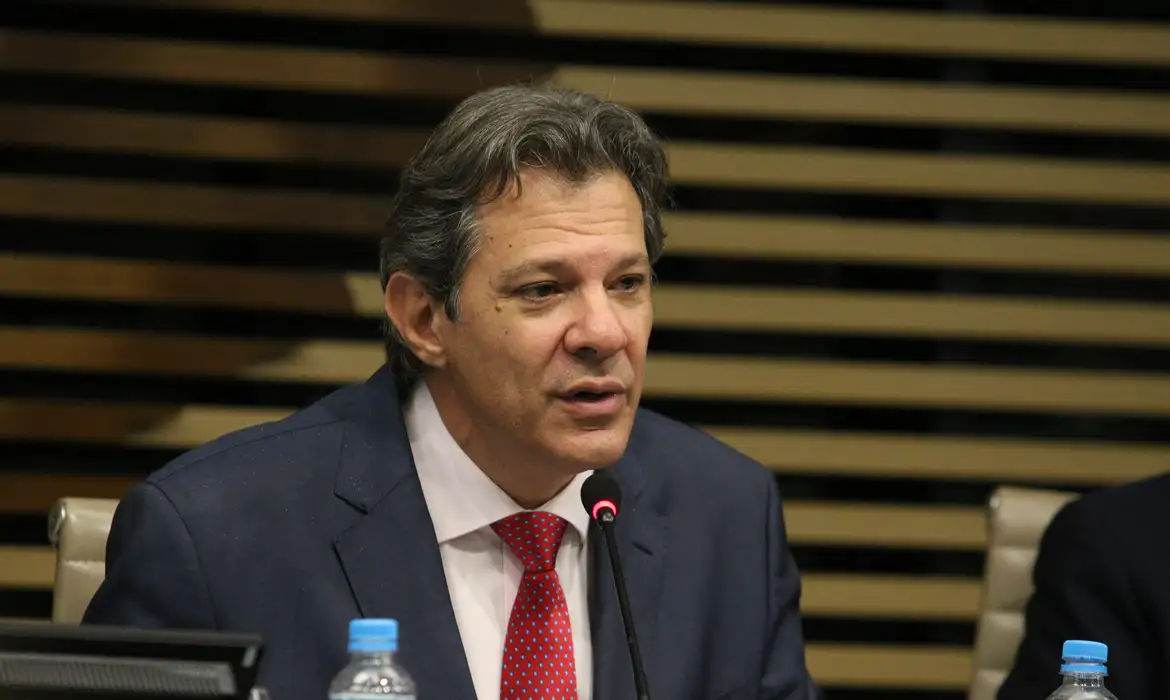 Fernando Haddad falando em microfone com expressão séria, sem olhar pra câmera
