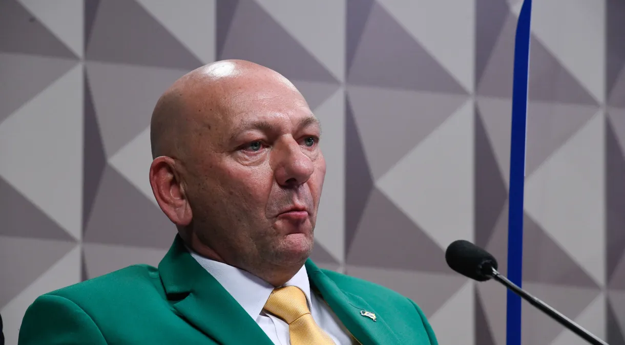 Luciano Hang de terno verde e gravata amarela, com camisa branca, fazendo careta sem olhar para a câmera