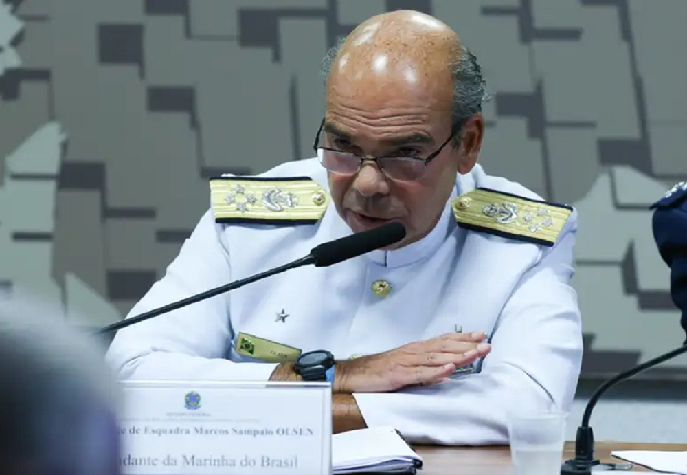 Marcos Olsen fardado, de óculos, falando em microfone com expressão séria