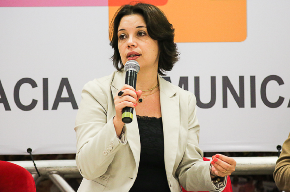 Renata Mielli falando em microfone e olhando para o lado com expressão séria