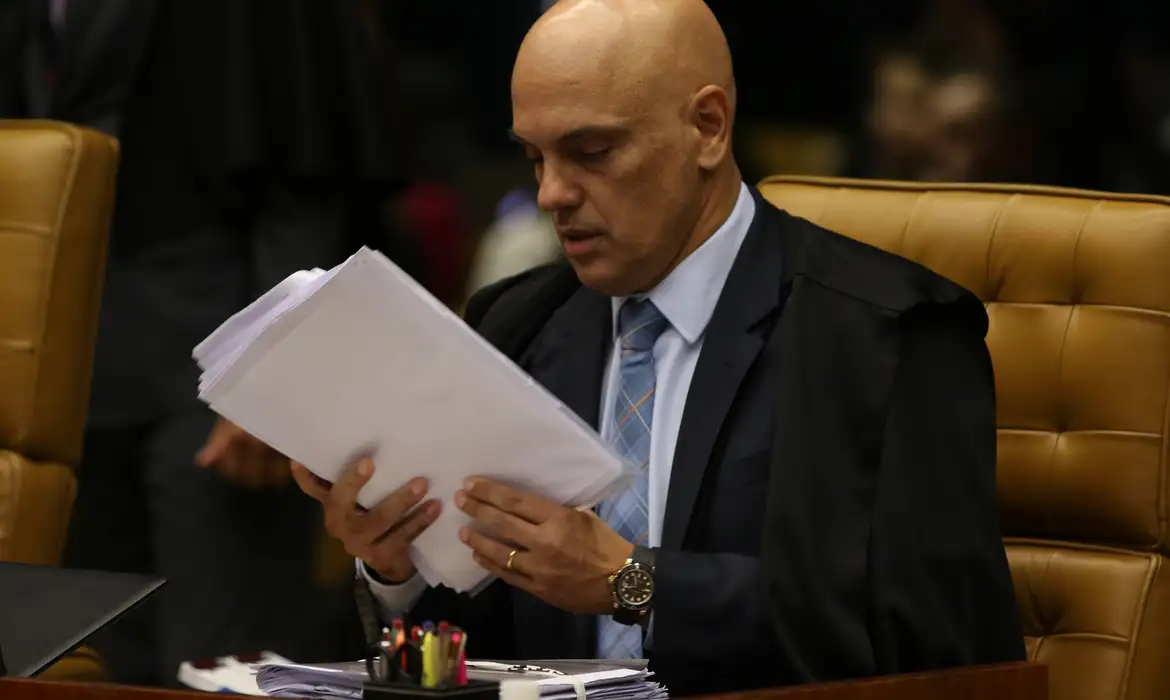Alexandre de Moraes com expressão séria, de cabeça baixa, lendo papeis