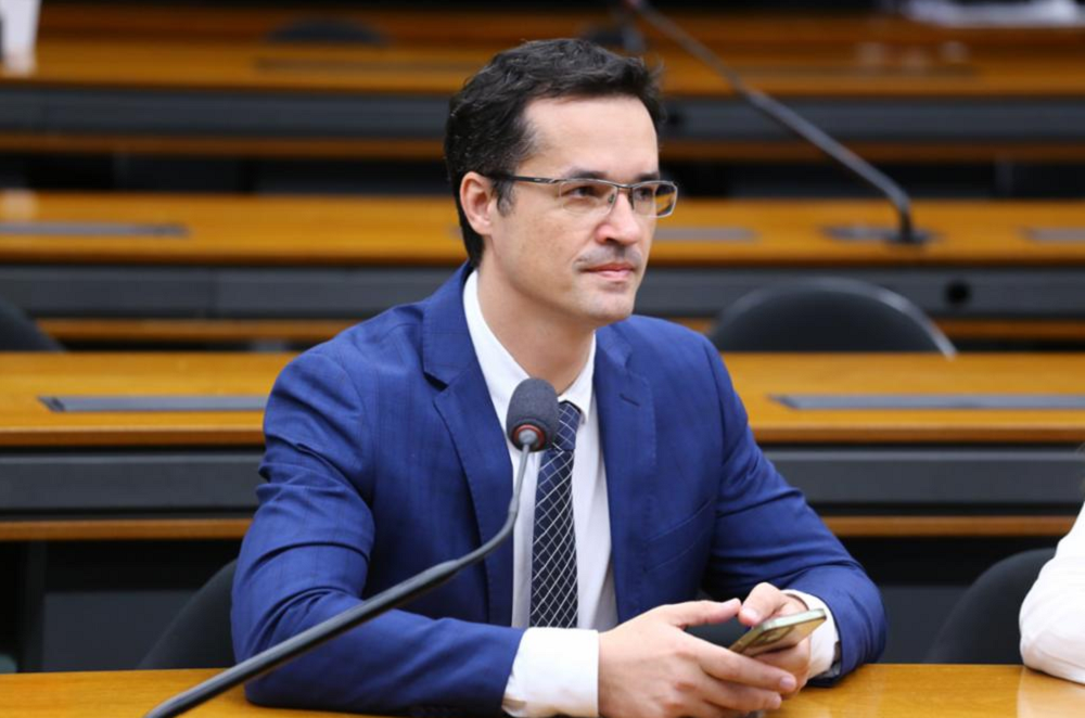 Deltan Dallagnol de roupa azul escura e óculos, sentado, sério, sem olhar para a câmera
