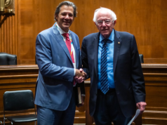 Fernando Haddad e Bernie Sanders sorridentes em aperto de mão, olhando para a câmera, ambos em pé