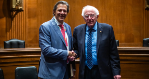 Fernando Haddad e Bernie Sanders sorridentes em aperto de mão, olhando para a câmera, ambos em pé