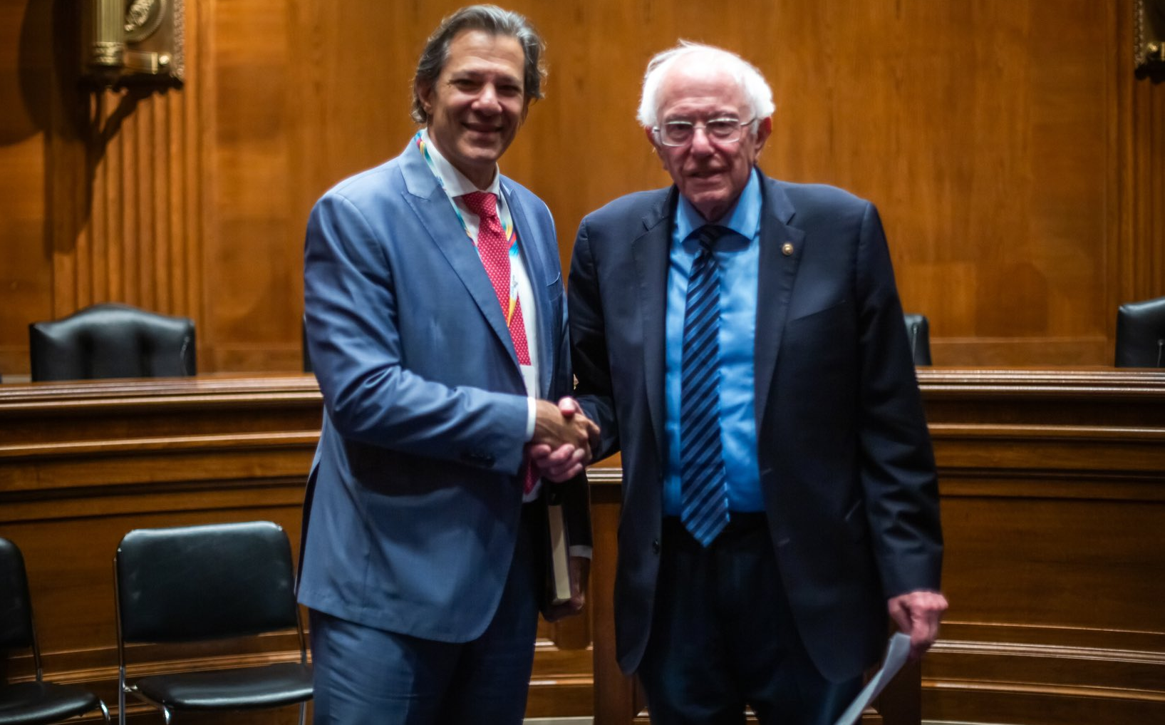 Fernando Haddad e Bernie Sanders sorridentes em aperto de mão, olhando para a câmera, ambos em pé