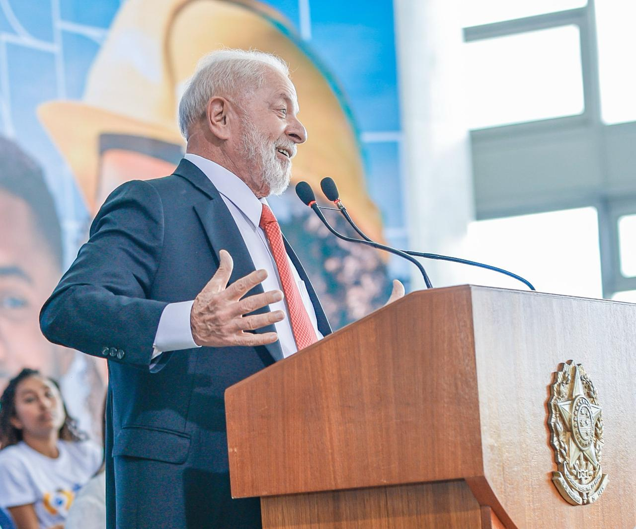 Lula de perfil, falando em microfone e gesticulando, sorrindo
