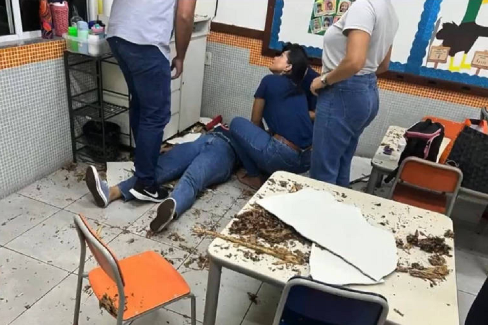 Professora caída em chão de sala de aula cercada de pessoas