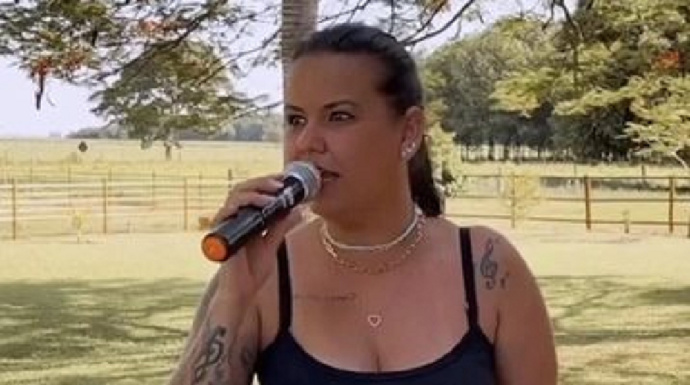 Nanda Ferraz cantando em microfone, de camiseta preta e rabo de cavalo, olhando para o lado