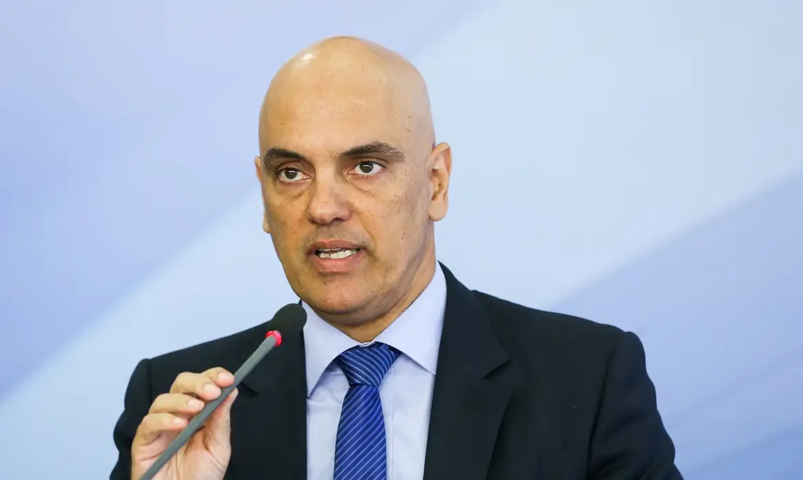 Alexandre de Moraes em fundo azul, falando em microfone, de terno e gravata, com expressão séria