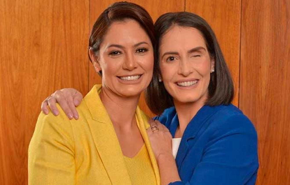 Michelle Bolsonaro e Amália Barros posando para foto abraçadas e sorrindo, a primeira de amarelo e a segunda de azul